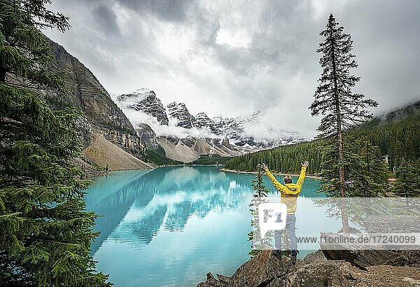 Young man stretching his arms in the air  cloudy mountain peaks  reflection in turquoise glacial lake  Moraine Lake  Valley of the Ten Peaks  Rocky Mountains  Banff National Park  Province of Alberta  Canada  North America
