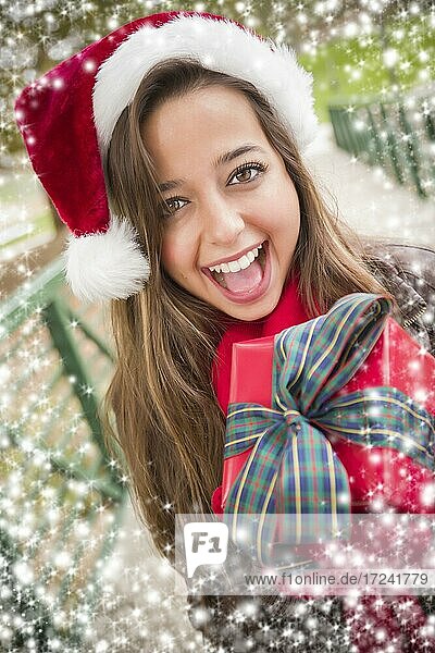 Hübsche festlich gekleidete lächelnde Frau trägt einen Weihnachtsmann Hut eingewickelt Geschenk draußen mit Schneeflocken