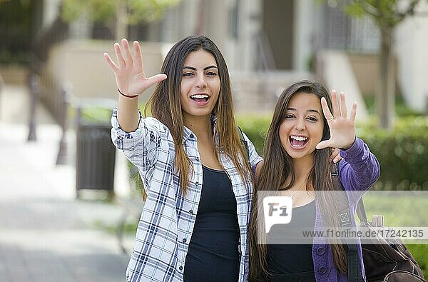Porträt von zwei attraktiven multiethnischen Studentinnen winken und tragen Rucksäcke auf dem Schulcampus