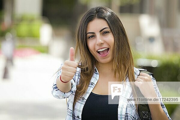 Attractive mixed race female student carrying backpack on school campus with thumbs up