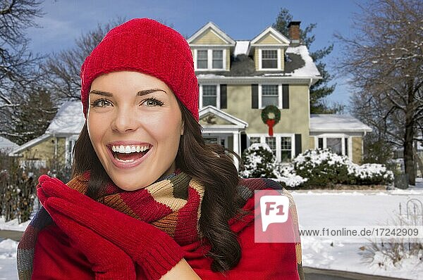 Lächelnde gemischtrassige Frau in Winterkleidung vor einem dekorierten Haus im Schnee