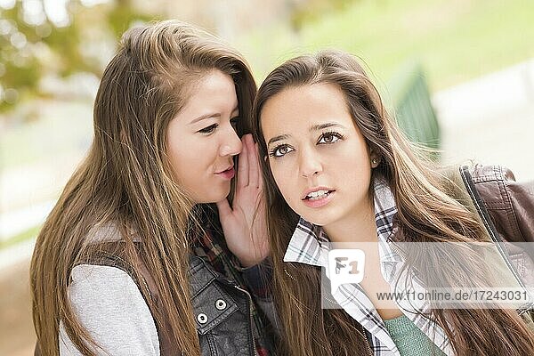Two attractive mixed-race woman with backpacks whispering secrets outside