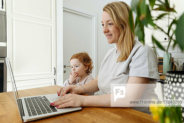 Lächelnde Unternehmerin  die mit ihrer Tochter am Tisch in der Küche an einem Laptop arbeitet