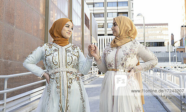 Freundinnen mit Hijab und Kleid lächelnd  während sie auf einer Brücke die Hände halten