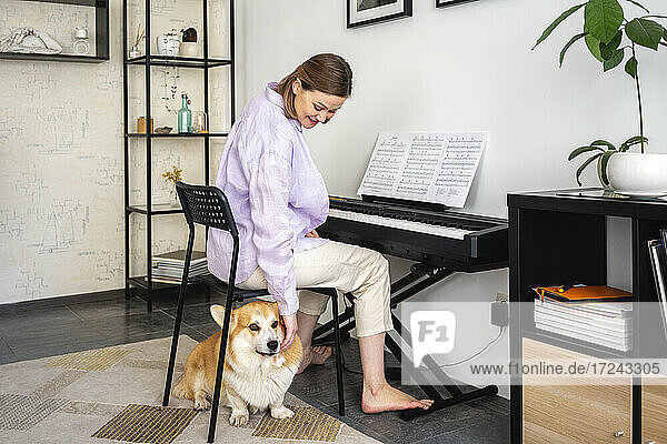 Lächelnde Frau  die ihren Hund streichelt  während sie im Wohnzimmer Klavier lernt
