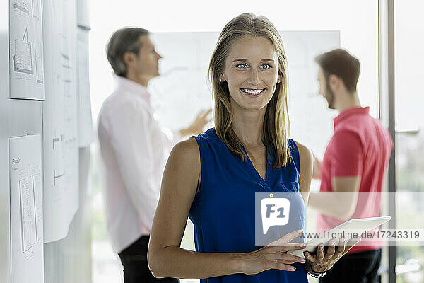 Young businesswoman holding digital tablet with male colleagues in background at office