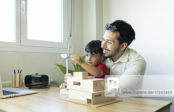 Männlicher Architekt lächelt  während sein Sohn mit dem Modell einer Windkraftanlage im heimischen Büro spielt
