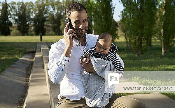 Lächelnder Mann  der mit seinem Handy telefoniert  während er einen kleinen Jungen im Park trägt