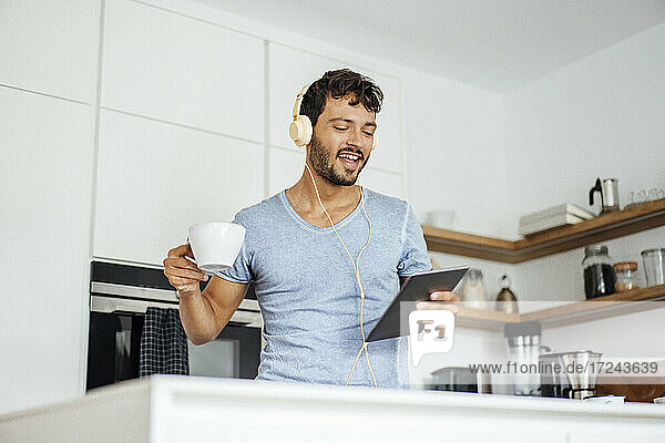 Junger Mann hält Kaffeetasse und Tablet in der Hand  während er in der Küche über Kopfhörer Musik hört