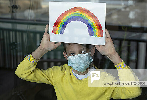 Junge mit Regenbogen auf Papier  der während der Pandemie eine Schutzmaske trägt  gesehen durch Glas