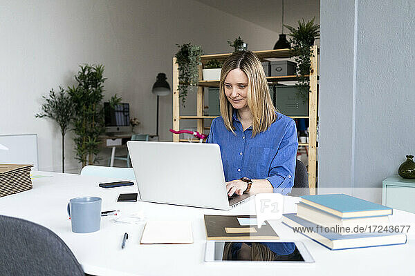 Lächelnde Geschäftsfrau mit Büchern auf dem Schreibtisch und Laptop im Büro