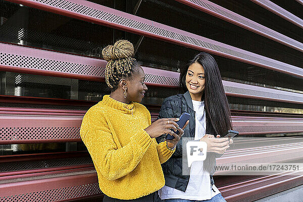 Multi-ethnische Freundinnen  die sich gegenseitig ansehen und ihr Smartphone an die Wand halten