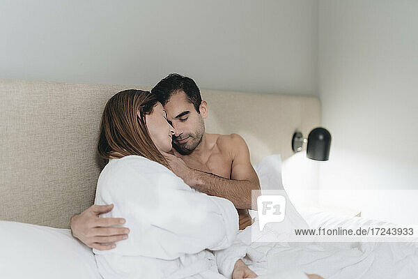 Mann ohne Hemd umarmt Frau  die auf dem Bett im Hotelzimmer sitzt