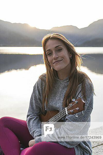 Happy woman with ukulele sitting at lakeshore