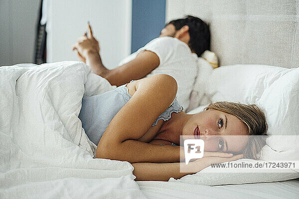 Nachdenkliche junge Frau  die sich hinlegt  während der Mann im Schlafzimmer ein Mobiltelefon benutzt