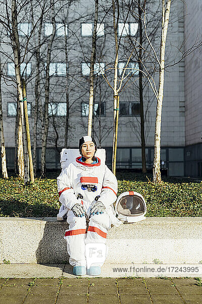 Young female astronaut in space suit sitting on retaining wall during sunny day