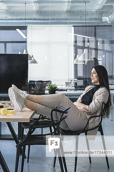 Lächelnde Geschäftsfrau mit hochgelegten Füßen  die wegschaut  während sie am Schreibtisch in einem Coworking-Büro sitzt