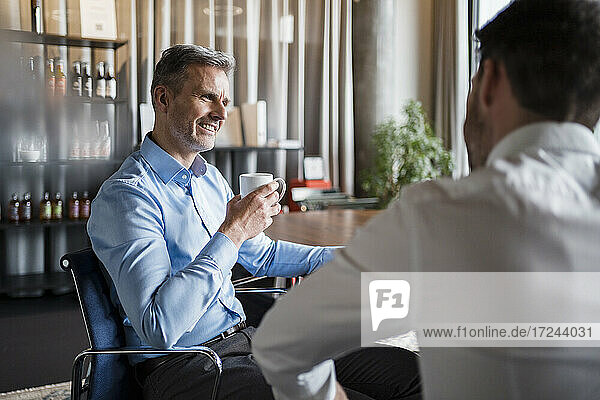 Älterer Geschäftsmann mit Kaffeetasse im Gespräch mit einem männlichen Kollegen am Arbeitsplatz
