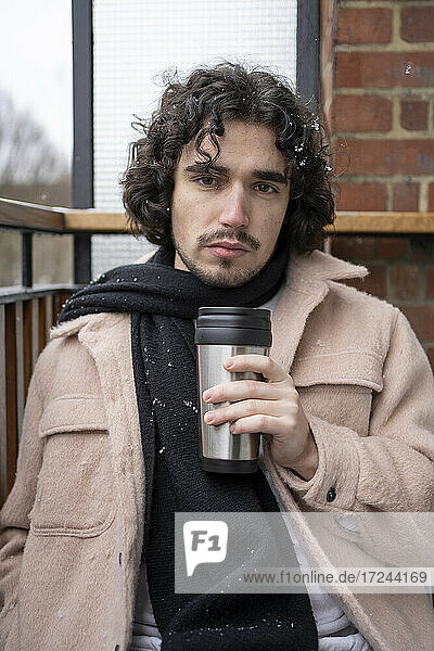 Young man staring while holding insulated drink container at balcony