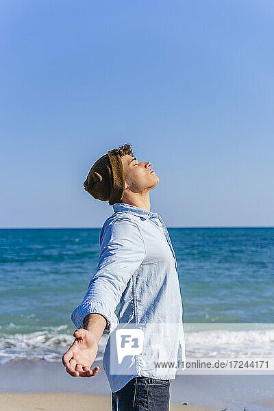 Junger Mann steht mit ausgestreckten Armen am Strand an einem sonnigen Tag