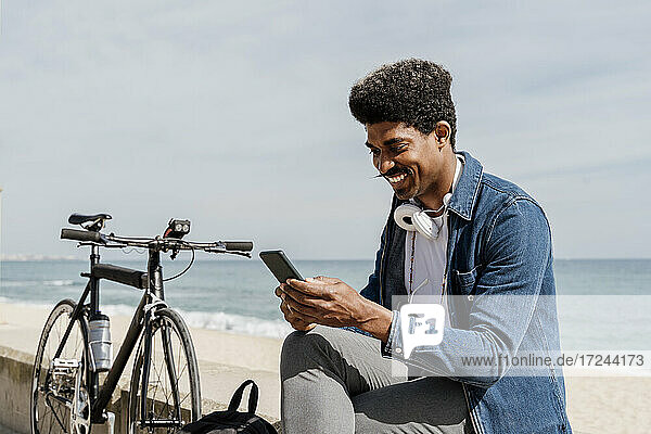 Lächelnder Mann mit Kopfhörern  der ein Mobiltelefon benutzt  während er an einem sonnigen Tag auf einer Stützmauer sitzt