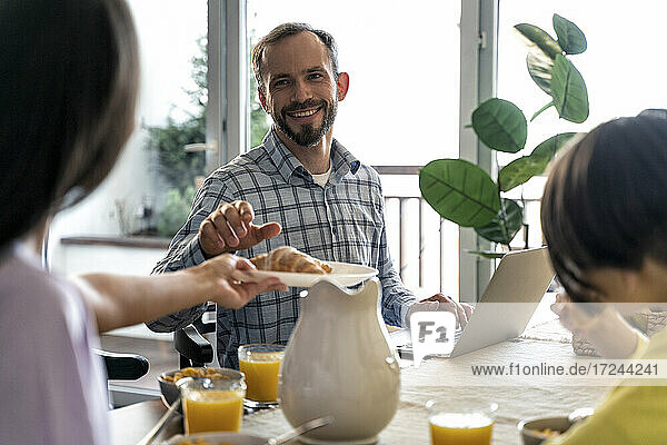 Lächelnder Mann mit Laptop sieht Frau beim Frühstück zu Hause an