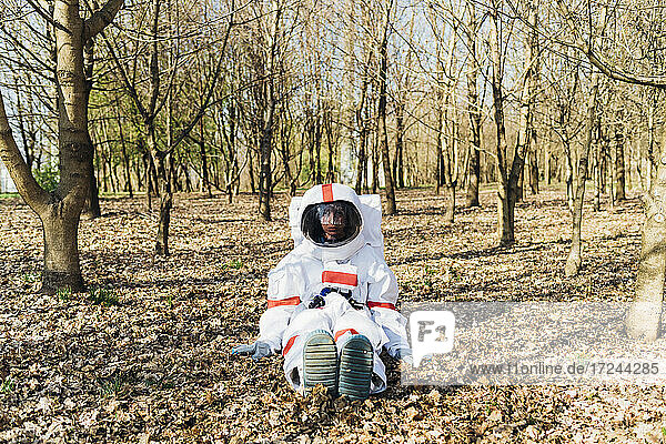 Female explorer wearing space suit and helmet sitting in forest
