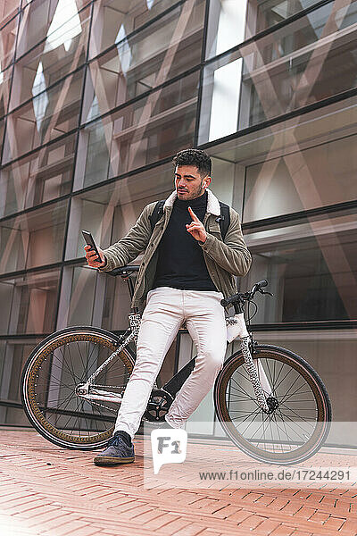 Männlicher Fachmann gestikuliert  während er ein Selfie macht und sich auf ein Fahrrad neben einem Bürogebäude lehnt