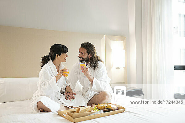 Paar in Bademänteln trinkt Saft auf dem Bett einer Hotelsuite