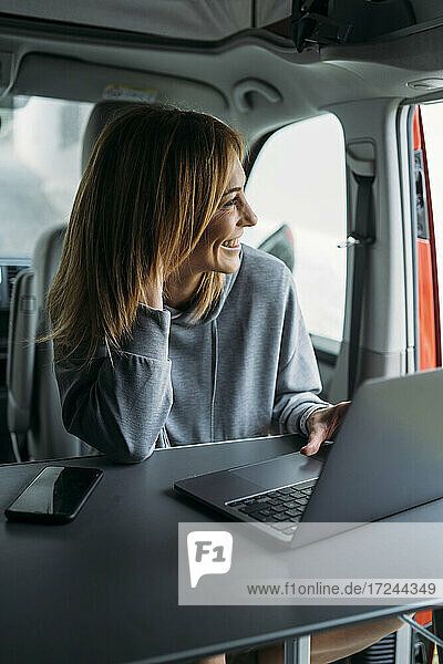 Female freelancer looking away while sitting with laptop and smart phone in van