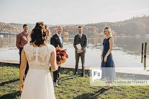 Bräutigam stehend mit Hochzeitsgästen mit Blick auf die Braut vor dem See auf Hochzeitsfeier