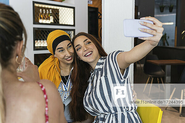 Lächelnde junge Frau macht ein Selfie mit einer kopftuchtragenden Freundin in einer Bar