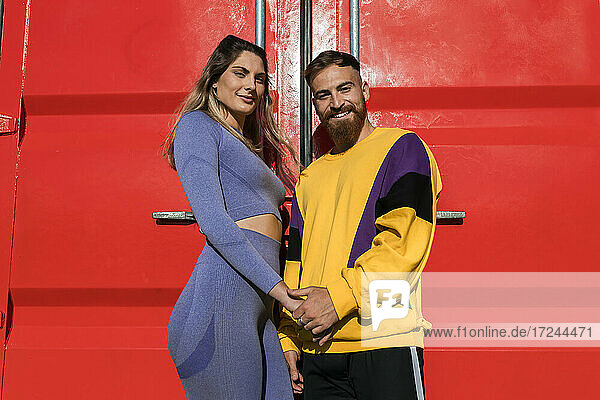 Lächelndes Paar hält sich an den Händen  während es vor einem roten Container steht