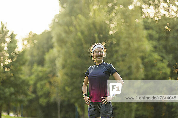 Lächelnde Frau mit Händen auf der Hüfte in einem öffentlichen Park stehend