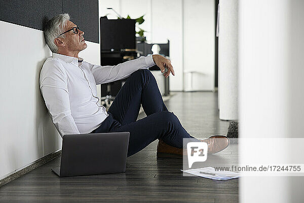 Müder Geschäftsmann sitzt mit Laptop auf dem Boden im Büro