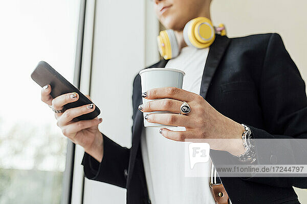 Geschäftsfrau  die eine Kaffeetasse hält und ein Mobiltelefon im Büro benutzt