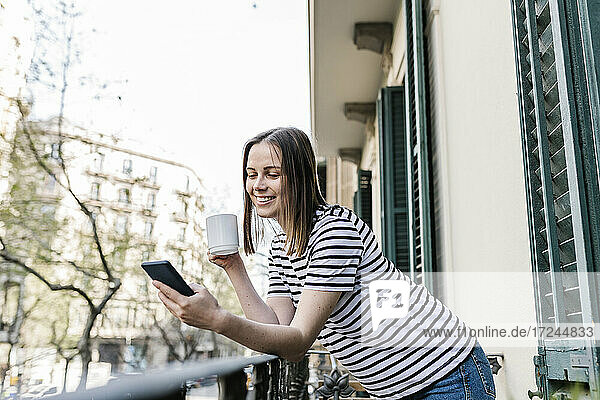 Lächelnde Frau mit Kaffeetasse  die ein Mobiltelefon benutzt  während sie sich auf einem Balkon an ein Geländer lehnt