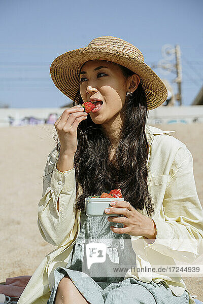 Schöne Frau mit Hut isst Erdbeeren  während sie am Strand an einem sonnigen Tag sitzt