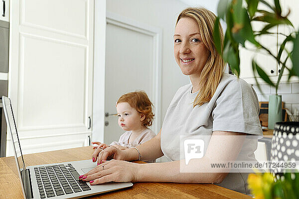 Smiling businesswoman using laptop by daughter at kitchen