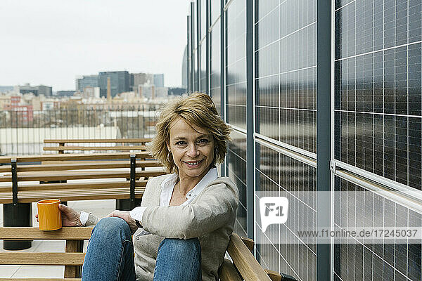 Lächelnde Geschäftsfrau mit Kaffeetasse an der Wand sitzend auf der Terrasse