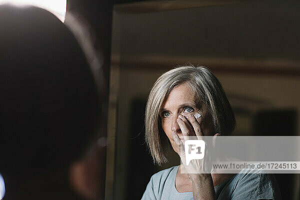 Kurzhaarige Frau schaut in den Spiegel  während sie zu Hause Gesichtscreme aufträgt