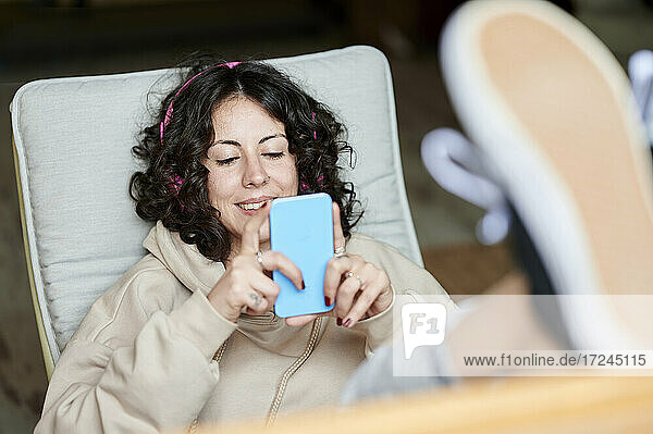 Lächelnde Frau mit hochgelegten Füßen  die zu Hause ein Mobiltelefon benutzt