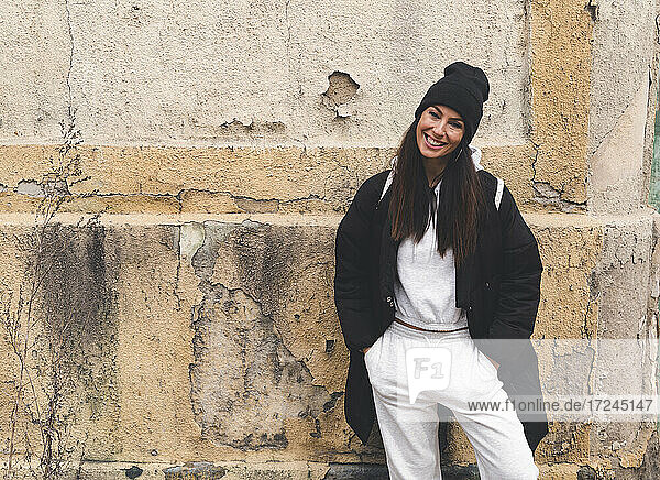 Smiling woman standing with hands in pockets in front of wall