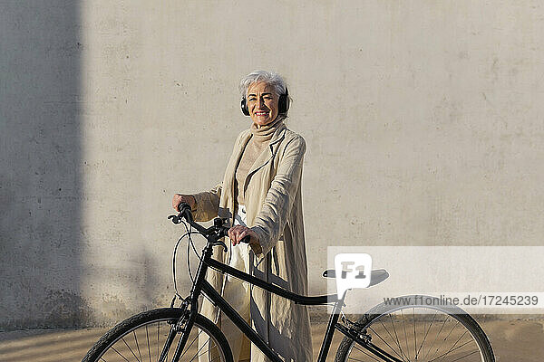 Lächelnde reife Frau mit Kopfhörern  stehend mit Fahrrad