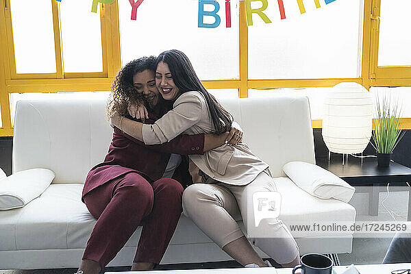 Smiling businesswomen embracing on sofa during birthday celebration at coworking office