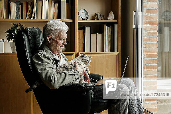 Senior man with laptop holding cat while sitting on chair at home