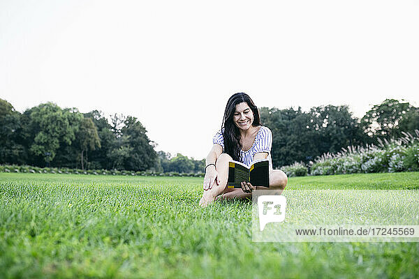 Lächelnde Frau liest ein Buch  während sie im Gras sitzt