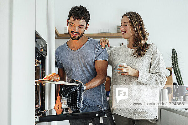 Lächelnde Frau sieht ihren Freund an  der ein gebackenes Croissant aus dem Ofen in der Küche nimmt