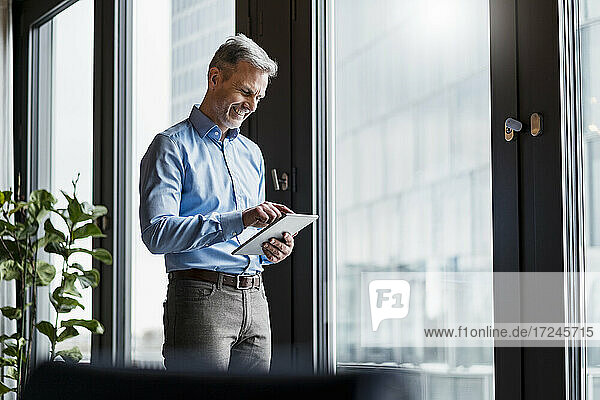 Smiling mature businessman working on digital tablet by window at work place