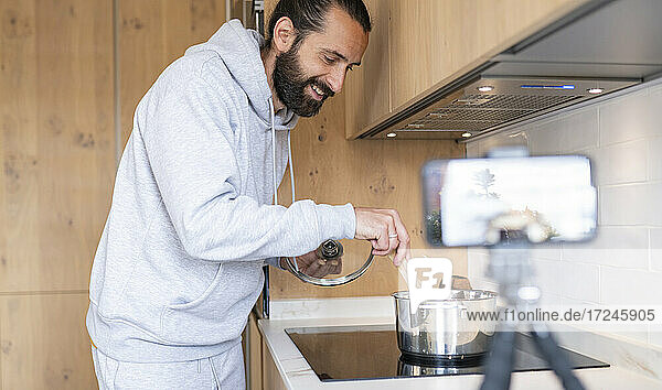 Smiling male influencer vlogging while preparing food in kitchen at home